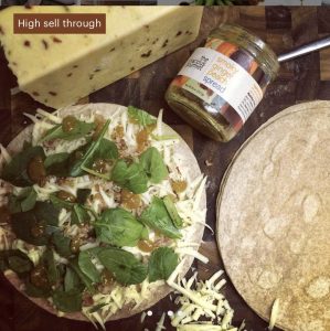 Tortilla with spinach, shredded cheese, and chutney next to a block of cheese, another tortilla, and a jar of spread.