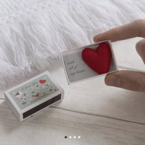 A hand holds an open matchbox with a red felt heart and a note reading "with all of my heart" next to a closed box.