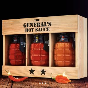 Three grenade-shaped bottles of hot sauce in a wooden box labeled "The General's Hot Sauce," with peppers in front.
