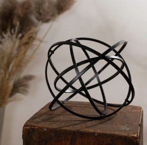A black metal geometric orb sculpture sits on a rustic wooden block with dried plants in the background.