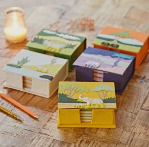 Five decorative boxes with animal and landscape designs are arranged on a wooden surface near a candle and pencils.