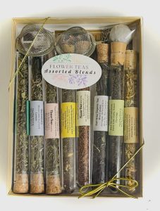 Box of assorted flower teas in glass tubes with corks, tea infusers, and labeled tags, tied with a gold ribbon.
