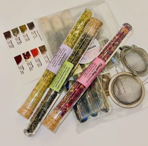 Three glass tubes of loose leaf tea blends with cork stoppers next to metal tea infusers and a tea blend guide card.