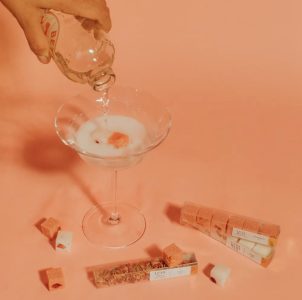 A hand pours liquid into a cocktail glass with cubes and small boxes of flavored cubes scattered on a peach-colored surface.