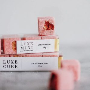 Two boxes of Luxe Cube strawberry champagne cubes with one partially eaten cube on top and one cube in the foreground.