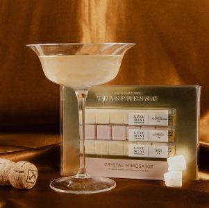 A coupe glass with a mimosa, a cork, sugar cubes, and a Teaspressa Crystal Mimosa Kit box on a brown satin surface.