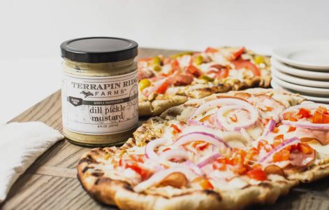 A jar of dill pickle mustard next to flatbread pizzas topped with onions, tomatoes, and cheese on a wooden board.