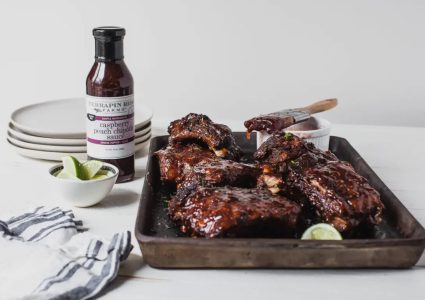 A tray of glazed ribs sits beside lime wedges, plates, and a bottle of raspberry peach chipotle sauce on a white table.