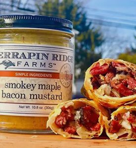 A jar of Terrapin Ridge Farms smokey maple bacon mustard next to sliced wraps filled with meat and vegetables.