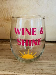 A clear glass with "WINE & SHINE" in pink letters and a yellow sun graphic at the bottom front.