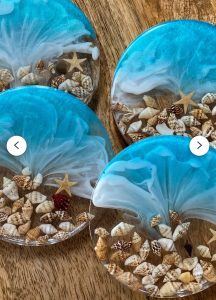 Four round resin coasters with blue, white, and sand designs, decorated with small seashells and starfish on a wooden surface.