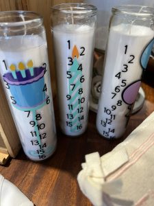 Three white pillar candles in tall glass holders, each decorated with numbers and birthday-themed illustrations.