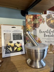 A slate cheese marker set, a book titled "Beautiful Boards," and a metal mortar and pestle displayed on a wooden shelf.