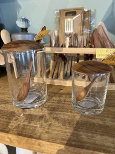 Two glass jars with wooden lids and matching wooden spoons are displayed on a wooden shelf in a kitchenware store.