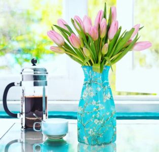 A blue vase with pink tulips next to a French press and a blue cup and saucer on a glass table by a window.