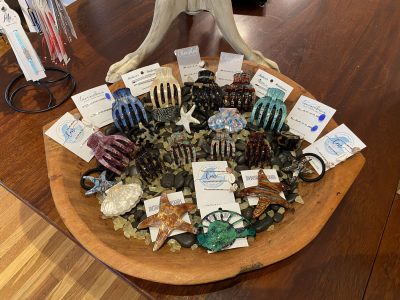 A wooden bowl displays assorted hair claws in various colors and designs, arranged with decorative rocks and small cards.