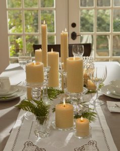 A dining table decorated with lit pillar candles, glass holders, ferns, and formal place settings near a bright window.