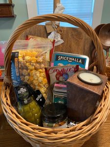 A wicker basket filled with snacks, seasoning tins, a candle, jarred pickles, and other gourmet food items.