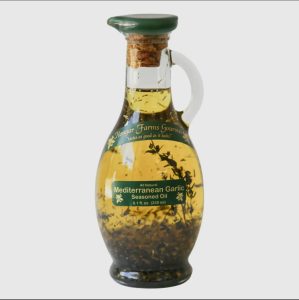 A glass bottle of Mangar Farms Mediterranean Garlic Seasoned Oil with herbs and garlic visible inside.