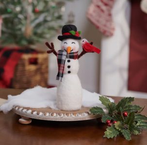 A felt snowman figure with a scarf, hat, and red bird stands on a wooden tray with holiday decor in the background.