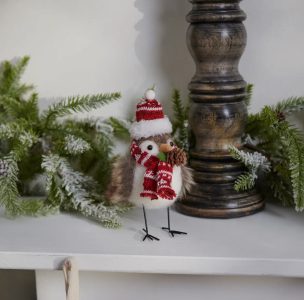 A decorative bird figurine wearing a red knit hat and scarf stands on a shelf with greenery and a candle holder.