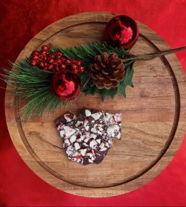 A piece of peppermint bark on a round wooden tray with pine, red ornaments, and a pine cone on a red background.