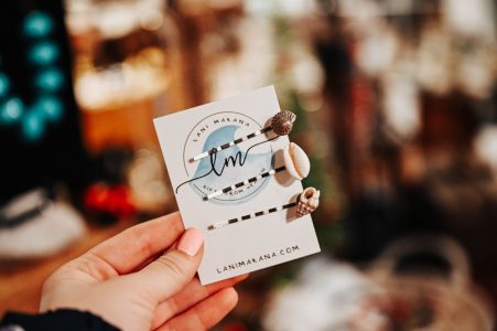 A hand holds a card displaying three bobby pins adorned with seashells, with branding text and website visible.