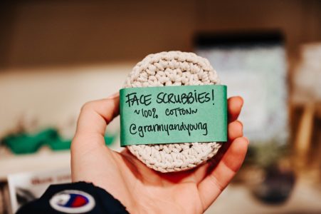 A hand holds a crocheted cotton face scrubbie labeled “FACE SCRUBBIES! ~100% Cotton @grammyandyoung.”.