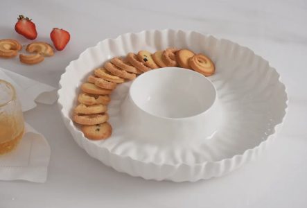 A white ceramic serving dish with assorted cookies around a central bowl, next to strawberries and a glass of juice.