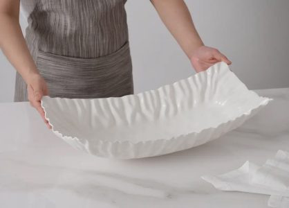 Person in a gray apron holds a large, white, wavy-edged ceramic platter on a white marble surface.