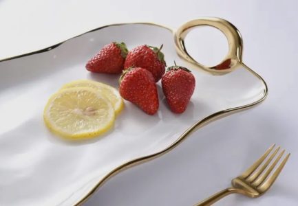A white and gold plate with three strawberries and two lemon slices next to a gold fork on a white surface.