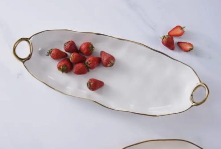 A white oval platter with gold handles holds strawberries; a few strawberry halves are placed beside the platter.
