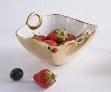 A gold and white bowl holding strawberries, with an extra strawberry and a grape on the white surface beside it.