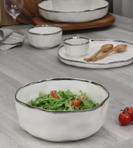 White ceramic bowl with silver rim filled with arugula and cherry tomatoes on a gray table with dishware in the background.