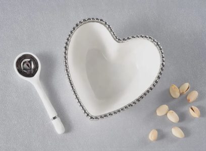 A white heart-shaped bowl, a white ceramic spoon, and several pistachio nuts on a gray surface.