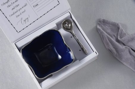 A gift box containing a dark blue square bowl and a metal spoon, next to a gray cloth on a gray surface.