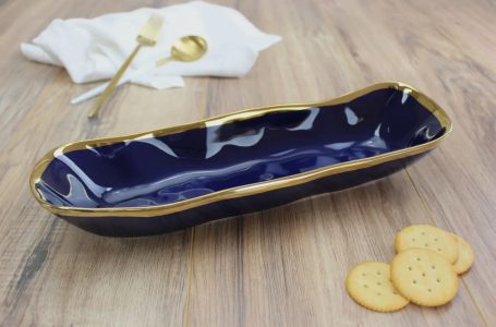 Navy blue ceramic serving dish with gold trim on a wooden surface, next to crackers and cutlery.