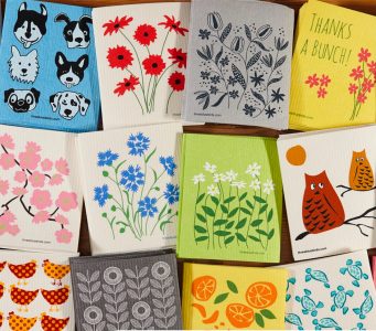 A variety of colorful dishcloths with floral, animal, and fruit designs arranged in a grid pattern on a wooden surface.