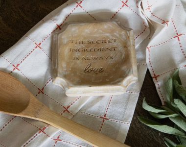 A wooden plaque reads "The secret ingredient is always love" on a white towel with a wooden spoon and herbs nearby.