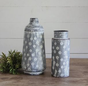 Two metal vases with white daisy patterns sit on a wooden surface next to a small green plant.