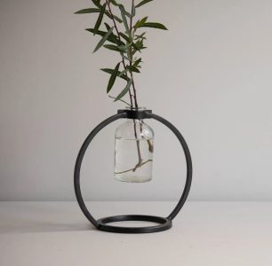 A glass vase with water and a leafy branch is suspended in a circular black metal stand on a plain surface.