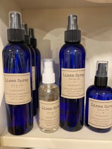 Several blue and one clear spray bottles labeled "Linen Spray" are displayed on a shelf, showing ingredient lists.