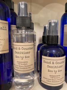 Clear and blue bottles labeled "Hand & Counter Sanitizer Zen by Zoe" arranged on a shelf.