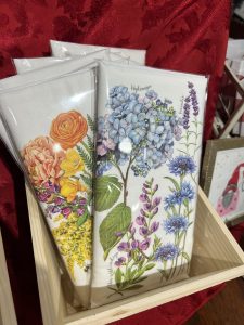 Two folded white tea towels with colorful floral designs are displayed upright in a wooden box.