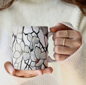 Person in a white sweater holding a mug with a white magnolia stained-glass design.