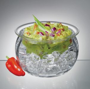 A glass bowl of guacamole with diced vegetables sits in a larger bowl filled with ice, next to a red chili pepper.