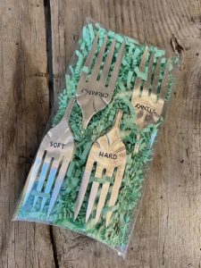 Four metal forks labeled "CRUMBLY," "STINKY," "SOFT," and "HARD" in a plastic bag on shredded green paper.