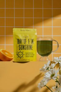 A yellow tea package, a glass of green tea, half an orange, and white daisies on a yellow tiled background.