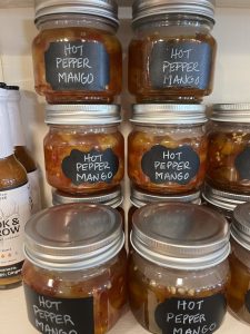Six glass jars labeled "Hot Pepper Mango" are stacked on a shelf, filled with a chunky orange mixture.