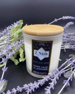 A white soy wax candle with a wooden lid labeled "White Sage & Lavender" surrounded by artificial lavender sprigs.